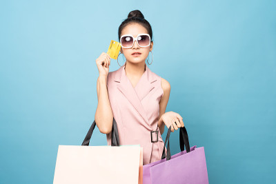 年轻漂亮可爱的亚洲女人拿着信用卡和购物袋的笑脸站在演播室。女孩戴着墨镜，穿着夏日休闲装。购物快乐，夏