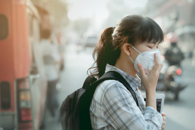 一个亚洲学生女孩戴着pm2.5口罩在一个充满灰尘和烟雾的城市里。