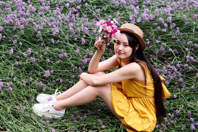 身着黄色服饰、头戴草帽的美丽女子坐在紫色的马鞭草花田里，一头乌黑长发的迷人中国姑娘在户外享受着她的闲