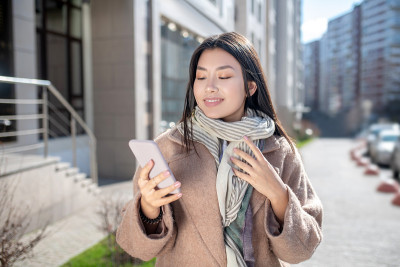 褐发女性，米色外套，站在外面看手机
