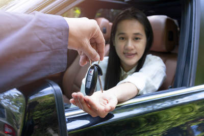 特写的经销商给钥匙的新主人的车。新车。汽车经销商给女人汽车钥匙，让她在乡村道路上试驾。软焦点微笑的妇