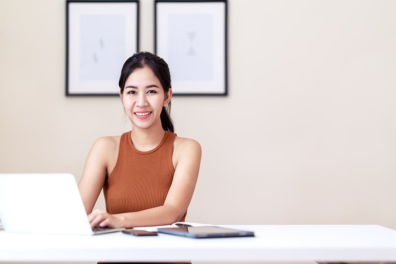 迷人的亚洲女企业家，快乐的学生或年轻的高管坐在白色的办公桌桌子上，穿着休闲的橙色背心裙微笑着看着相机