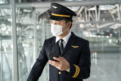 亚洲航空的飞行员戴着口罩，站在机场候机楼。年轻英俊的机长在COVID - 19大流行危机期间用手机聊