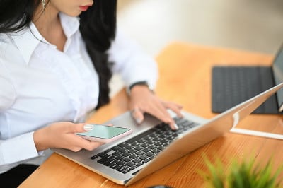 一名女性手持智能手机，在笔记本电脑键盘上打字。