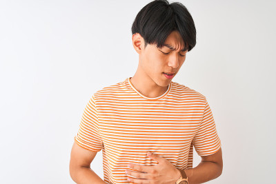 年轻的中国男子穿着休闲条纹t恤站在孤立的白色背景与手对胃，因为消化不良，痛苦的疾病感觉不舒服。疼痛的