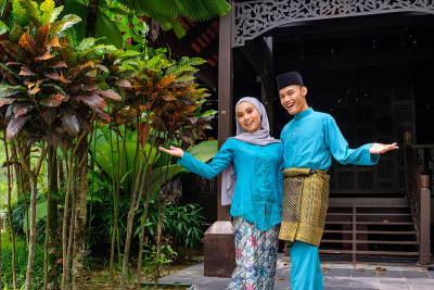 年轻的马来西亚穆斯林夫妇在传统服装在Aidilfitri庆典上显示欢迎欢迎的手势木制房子。