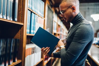 白种男学生站在旧公共图书馆的书架旁选择书籍阅读，侧视学术教授花时间在档案中选择文献以增加知识