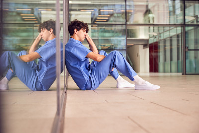 疲惫的年轻男医生穿着手术服坐在现代医院大楼靠墙