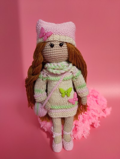 钩针娃娃女孩在针织连衣裙和帽子与蝴蝶在粉红色的背景。Amigurumi手工制作的玩具