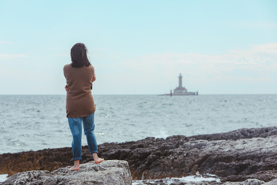 一个穿着湿牛仔裤的女人走在布满岩石的海滩上，背景是灯塔