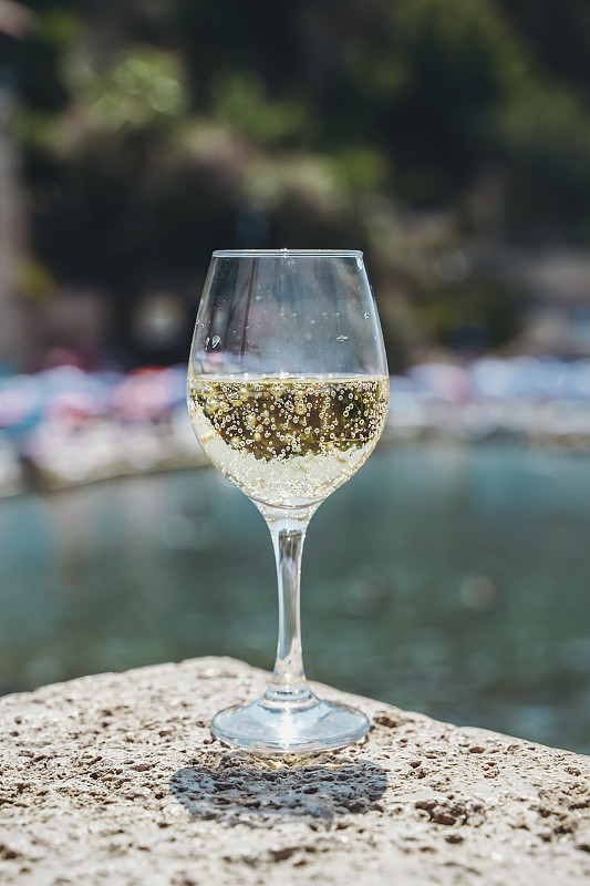 一杯白葡萄酒，映衬着地中海海滩和夏日阳光下的旅游小镇的大海。夏日，旅行，生活方式，放松，享受的概念。