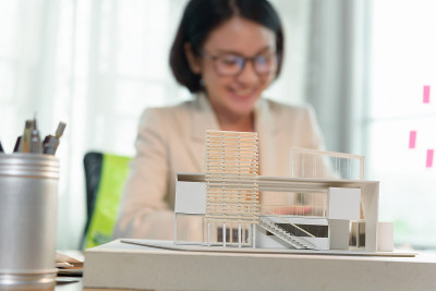 办公桌办公室的现代盒子屋模型，幸福的亚洲女性建筑师-设计师坐在工作背景中