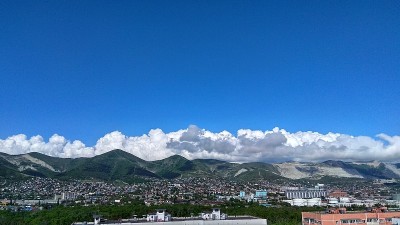 在蓝天下，青山后面密密麻麻、洁白蓬松的云，点缀着房屋，周围是一片绿色的森林。