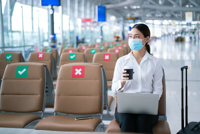 新常态与社会距离概念。2019冠状病毒疫情期间，亚洲年轻女性游客戴着口罩，用笔记本电脑搜索航班状态，