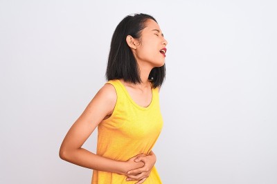 年轻的中国女子穿着黄色休闲t恤站在孤立的白色背景与手对胃，因为消化不良，痛苦的疾病感觉不舒服。疼痛的