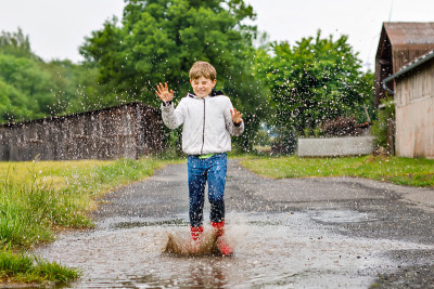 快乐的小男孩穿着红色雨靴，在雨夹雪和阴雨天行走。孩子在五颜六色的休闲衣服跳进水坑。在户外玩，健康的儿