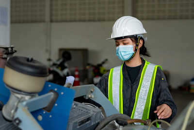 年轻的亚洲女工程师戴着医用口罩遮住嘴，戴着白色安全帽检查施工区内的大型地板切割机。