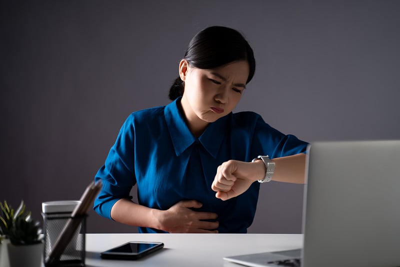 一名身穿蓝色衬衫的亚洲妇女坐在办公室里胃痛。孤立在白色背景上。