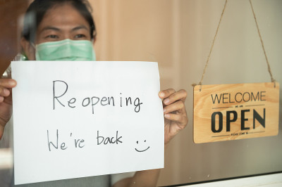 年轻的亚洲企业家咖啡店经理戴着外科口罩贴着重新开业的标志，宣布因冠状病毒爆发的商业危机而关闭的店铺重