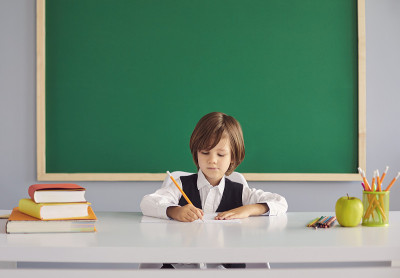 回到学校。一个长头发的有趣的小男孩在桌子旁的绿色黑板上写着什么