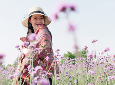 身穿黑色马鞭草背心、披着彩色披肩、戴着草帽的美丽女子，在紫色的马鞭草花海中摆姿势，一头乌黑长发的迷人