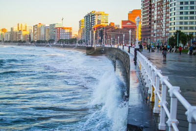 西班牙阿斯图里亚斯省希洪市圣洛伦佐海滩，海浪拍打着海滩的墙壁，背景是建筑物和散步道
