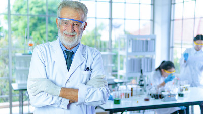 英俊的资深科学家男子穿着白色大褂站在室内实验室背景前微笑着积极。成功的面孔和情感的表达。