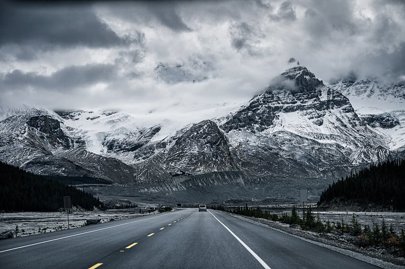 在阴暗的冰原公园道路上与多雪的岩石山脉的公路旅行