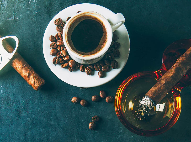 一杯咖啡，咖啡豆，烟灰缸和雪茄在黑暗的背景。