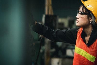 女工业工程师戴着白色的头盔，站在她身后的一个重工业工厂看工作在工业机械和检查安全系统安装在工厂。