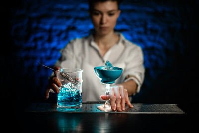 调酒师手持蓝色杯子和调酒杯与蓝色冷鸡尾酒站在吧台上