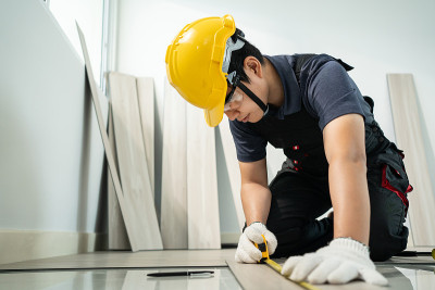 亚洲建筑工人在室内地板上安装强化板。工匠建筑工人或木匠男戴安全帽，用卷尺测量走廊上的拼花地板来翻新公
