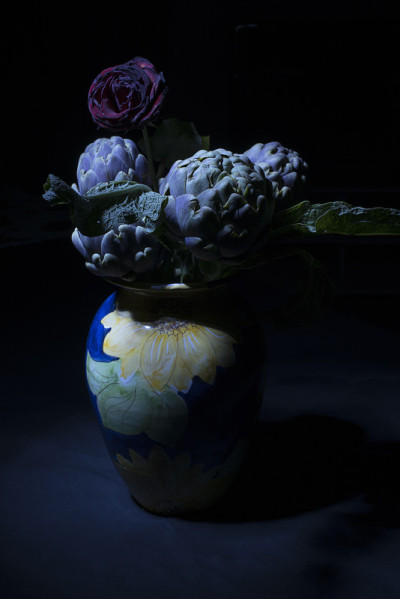 蓝色花瓶里装着一朵玫瑰和三枚洋蓟。夜景拍摄在一个手绘的带有花卉图案的蓝色陶瓷花瓶里。