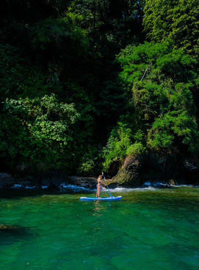 SUP直立桨板。金发女孩穿着泳衣在石头海滩附近的绿色海上划桨板上航行。