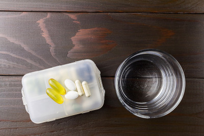 白色和黄色的氨基葡萄糖胶囊，omega-3钙片放在塑料盒上，玻璃与水放在木桌上，俯视图