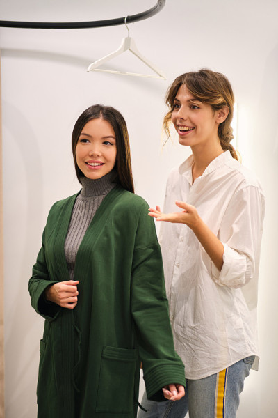 年轻的时尚顾问愉快地帮助在现代商店里美丽的亚洲女孩试穿开襟羊毛衫