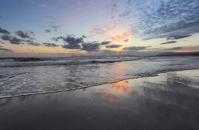 Agia Marina Beach，克里特岛，希腊。海岸边的岩石闪闪发光，日出反射在潮湿的沙子上。早
