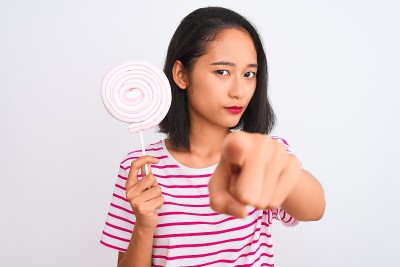 年轻美丽的中国女子站在孤立的白色背景下吃着棒棒糖，手指指着镜头和你，手势，正面自信的手势