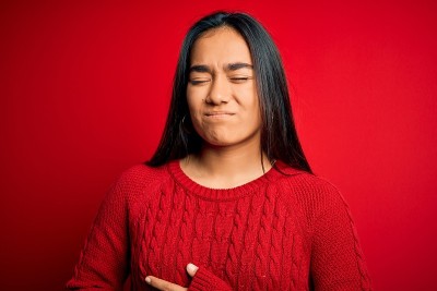 年轻美丽的亚洲女人穿着休闲毛衣站在孤立的红色背景与手对胃，因为消化不良，痛苦的疾病感觉不舒服。疼痛的
