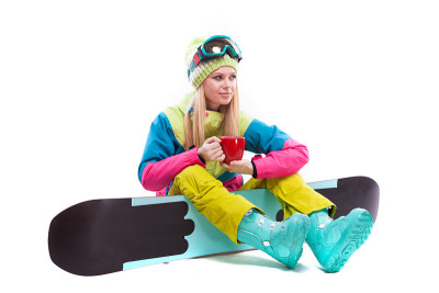 迷人的年轻女子穿着滑雪服和滑雪眼镜坐在滑雪板上，手持红色杯子