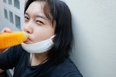 亚洲泰国妇女戴白布口罩以预防Covid-19或冠状病毒，喝瓶装橙汁以补充维生素c有助于提神。泰国的空