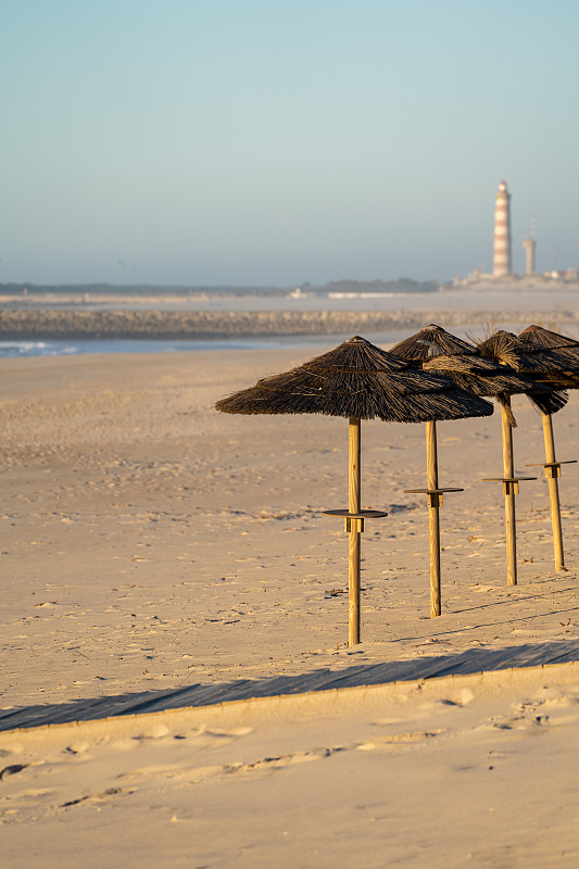 日落时分，海滩上的草棚撑着遮阳伞，普拉亚·达·巴拉灯塔(Lighthouse of Praia da