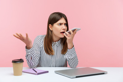 愤怒沮丧的商务女性在条纹衬衫留下语音信息，拿着手机靠近嘴坐在工作场所，给语音助手命令