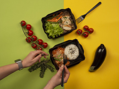 女人的手拿着叉子去拿外卖盒里的食物:米饭配酱鱼，鸡肉配荞麦，蔬菜和绿色蔬菜。