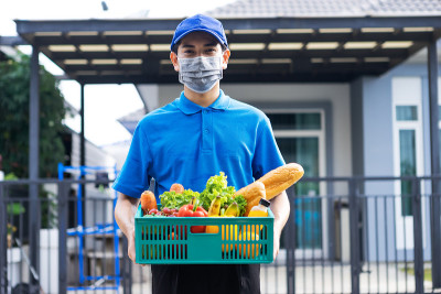 亚洲快递员身穿蓝色制服，戴着医用口罩，提供食品杂货服务，为客户提供新鲜食品，食品递送和杂货服务理念。