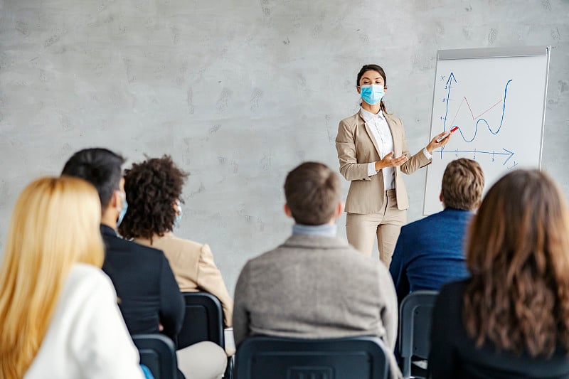 经济学家关于金融和加密货币的演讲。一个带着口罩的女商人站在一张图表旁边，指着它做一个关于经济的演讲。