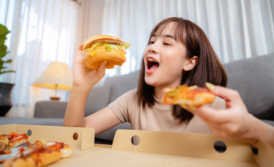 饥饿的年轻女人吃垃圾食品汉堡和披萨午餐订购送货上门度假。不健康饮食，肥胖风险。