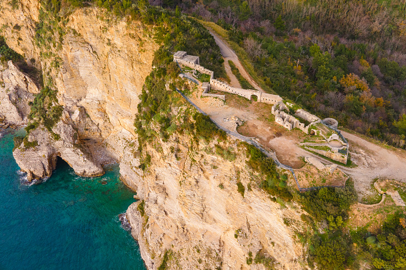 在黑山的Budva，一架带着相机的无人机在Mogren要塞的废墟上升起，它被森林和道路包围，坐落在被