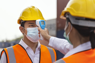 亚洲男性工人戴着口罩，在施工现场为预防新冠肺炎、新冠肺炎或冠状病毒大流行而进入工作之前，用数字体温计