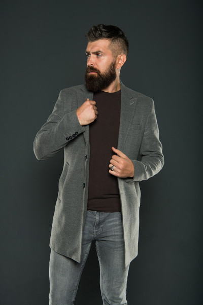 Beardy潮人。时髦的灰色背景。留胡子的潮人穿休闲夹克和牛仔裤。时髦的野蛮男人
